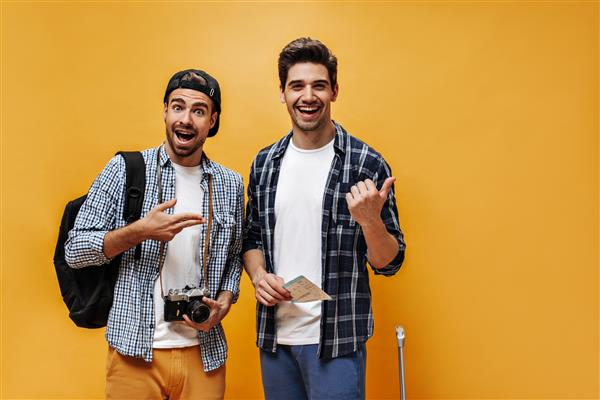 مسافران جوان شاد و باحال روی دیوار نارنجی شادی می کنند مردان شاد با پیراهن های چهارخانه با بلیط هواپیما و دوربین یکپارچهسازی با سیستمعامل ژست می گیرند