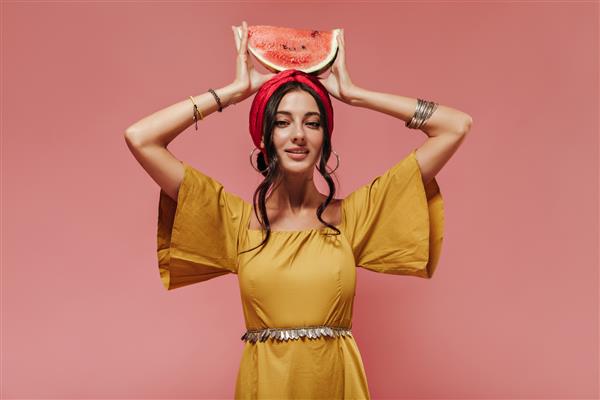 دختر هندی با موهای موج دار مشکی با هدبند و لباس های مد روز زرد که هندوانه را روی سرش روی دیوار صورتی نگه داشته است