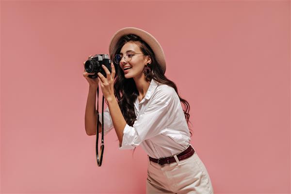 خانم شیک پوش فوق العاده با مدل موی شیک در گوشواره قهوه ای عینک شفاف و لباس سفید ژست و دوربین در دست