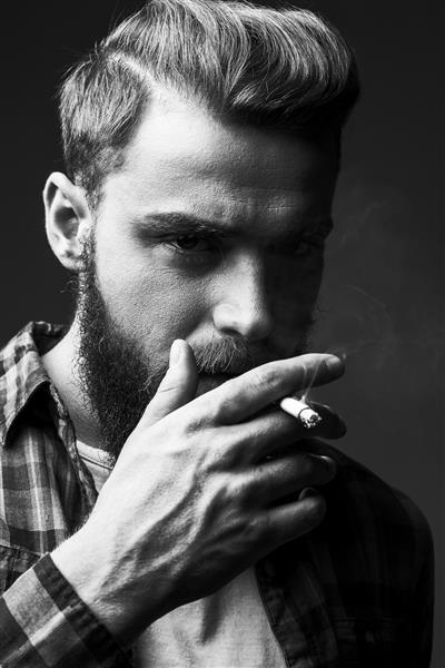مرد ریشو در حال سیگار کشیدن پرتره سیاه و سفید مرد جوان ریش دار خوش تیپ در حال کشیدن سیگار و نگاه کردن به دوربین در حالی که در پس زمینه خاکستری ایستاده است