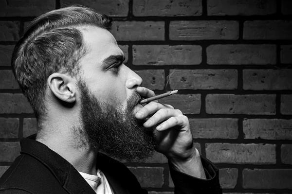 مردی که سیگار می کشد پرتره سیاه و سفید مرد جوان ریش دار خوش تیپ در حالی که در مقابل دیوار آجری ایستاده سیگار می کشد