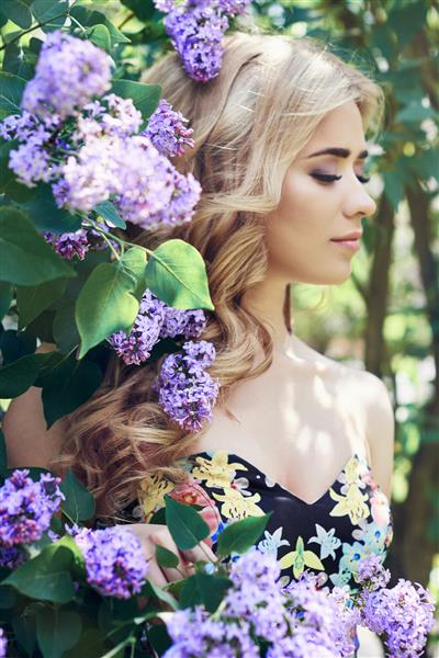 زن جوان زیبای مد در فضای باز که در تابستان با گلهای یاس بنفش احاطه شده است بهار شکوفه بوته یاس بنفش پرتره دختر بلوند
