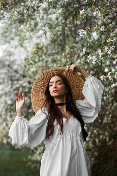 زنی با موهای قهوه ای با کلاه حصیری بزرگ و لباس سفید در پس زمینه درختان سفید شکوفه ژست می گیرد ظاهر رمانتیک زیبایی طبیعی پوست صورت تمیز