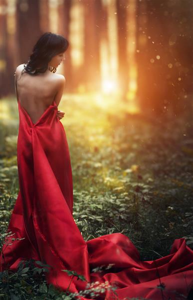 زنی با لباس قرمز بلند به تنهایی در جنگل تصویر افسانه ای و مرموز از یک دختر در یک جنگل تاریک در آفتاب عصر غروب آفتاب در جنگل شاهزاده خانم گم شد
