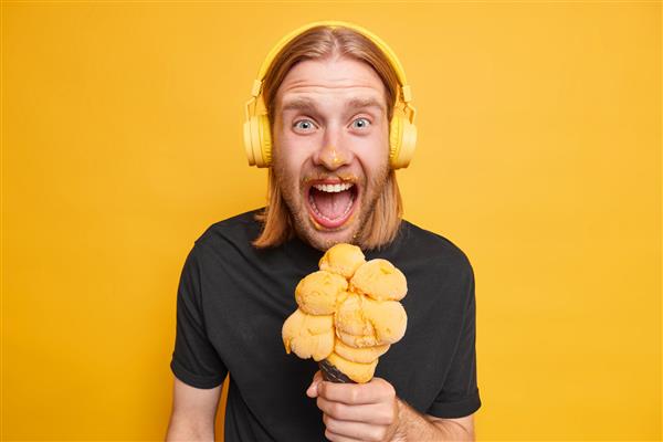 عکس استودیویی از مرد مو قرمز احساسی که بعد از خوردن بستنی مخروطی کثیف شده و از دسر تابستانی خوشمزه لذت می برد غذای خوشمزه را برای خودش تهیه می کند و آهنگ صوتی را با هدفون گوش می دهد