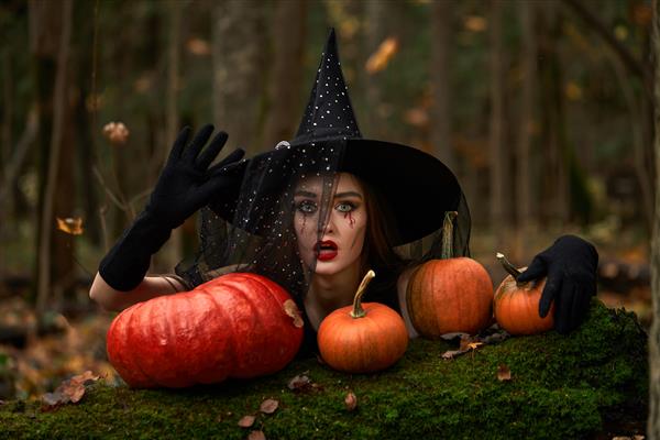 زن جوان با لباس مشکی با کلاه جادوگر و کدو تنبل نارنجی در جنگل مفهوم هالووین تم ترسناک