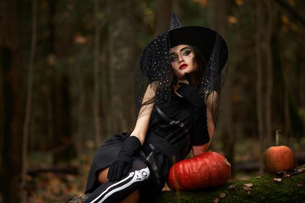 زن جوان با لباس مشکی با کلاه جادوگر و کدو تنبل نارنجی در جنگل مفهوم هالووین تم ترسناک