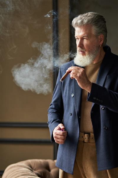 مرد میانسال وحشی شیک با ریش و در حالی که سیگار می کشد در اتاق زیر شیروانی ایستاده است و به سمتش نگاه می کند