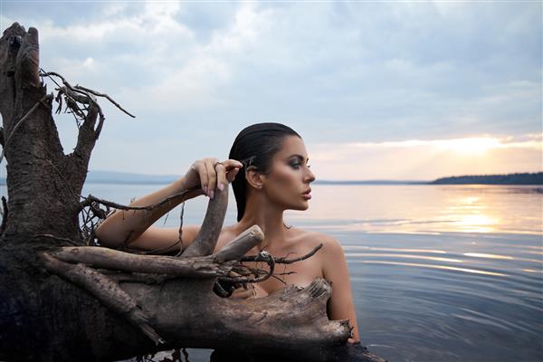 زن زیبای سبزه با لباس مشکی در دریاچه دریا در پس زمینه آسمان آبی ژست می گیرد زن زیبا موهای بلند و آرایش زیبایی زیبا روی صورتش