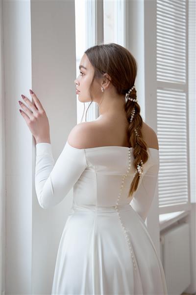 بلوند لاغر زیبا در آفتاب عصر با لباس سفید بلند در نزدیکی یک پنجره بزرگ آرایش و موی عالی برای عروس مجموعه ای جدید از لباس عروس