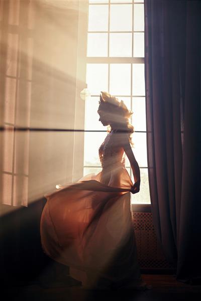 شبح یک دختر زن زیبا در نزدیکی پنجره در آفتاب صبحگاهی زن بلوند مد در زیر پرتوهای خورشید نور خورشید بر بدن زن می افتد نور جادوی پری در پنجره