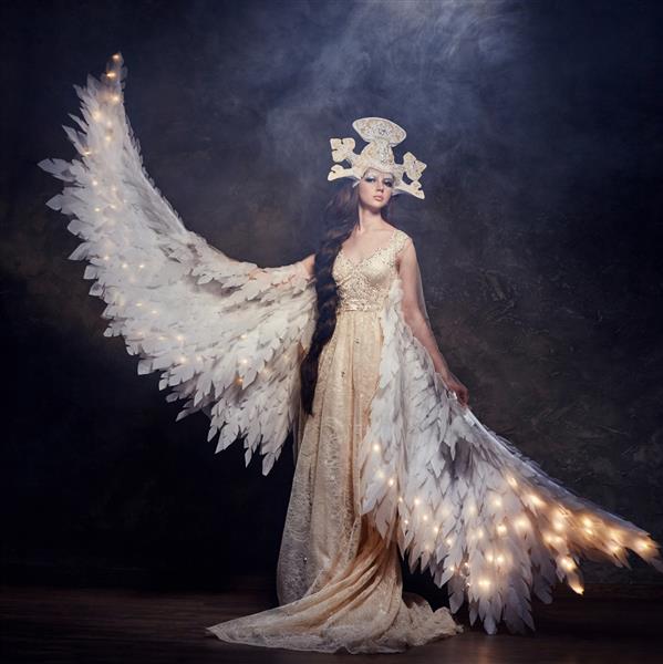 فرشته زن هنری با بال در یک لباس بلند مجلل و سرپیچ افسانه ای پرنده دختر با بال های درخشان که روی پس زمینه تیره ژست گرفته است