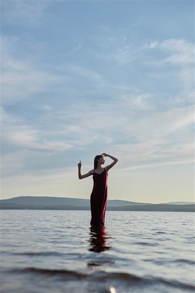 زن زیبا با لباس قرمز روشن در غروب آفتاب در آب دریاچه ایستاده است انعکاس دختر و آسمان در آب