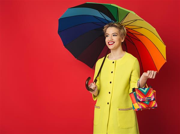 دختر مد شاد با کیف و چتر رنگین کمانی در پس زمینه قرمز زن جوان و شیک در استودیو فضای کپی