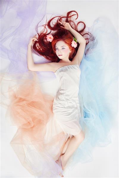 زن رمانتیک با موهای بلند و لباس ابری زنی که رویای آرایش روشن و اندام عالی دارد زن مو قرمز با لباس رنگی روشن روی زمین دراز کشیده است گل های زیبا در موهای زن
