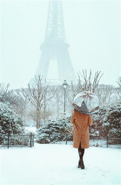 زن جوان با کت بژ زیر چتر در پاریس برفی زمستانی راه می‌رود