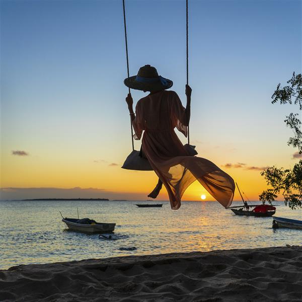 دختری زیبا با کلاه حصیری و پارئو در حال تاب خوردن بر روی تاب در ساحل هنگام غروب آفتاب جزیره زنگبار تانزانیا آفریقا مفهوم سفر و تعطیلات