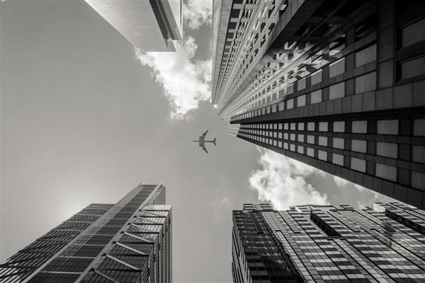 عکسی در مقیاس خاکستری با زاویه کم از یک هواپیما که در بالای ساختمان های مرتفع پرواز می کند