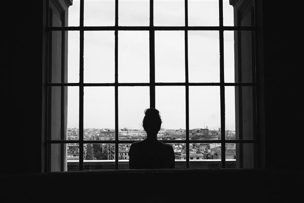 عکس سیاه و سفید از یک زن تنها که جلوی پنجره ها ایستاده و به ساختمان ها نگاه می کند