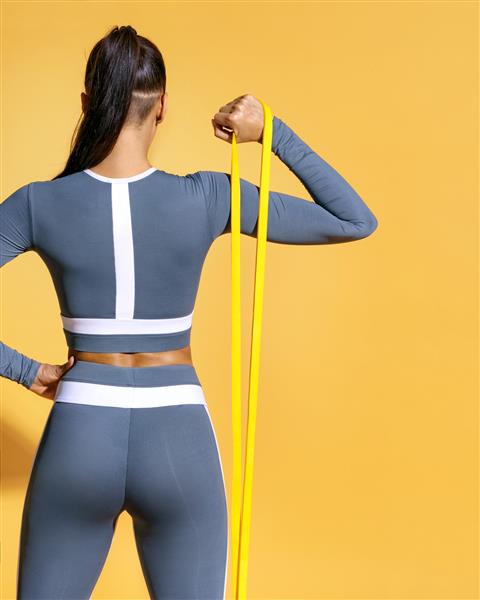 دختر تناسب اندام با باند مقاومتی تمریناتی را برای عضلات دست انجام می دهد عکس دختر لاتین با لباس ورزشی شیک در زمینه زرد قدرت و انگیزه نمای عقب