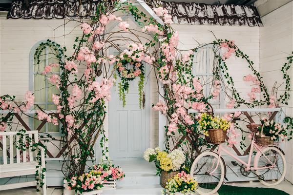 فضای داخلی یک آتلیه عکس روستایی نمای ساختمان سفید با ایوانی تزئین شده با گل های شکوفه سبدهایی با گل در کنار پله ها و یک دوچرخه صورتی