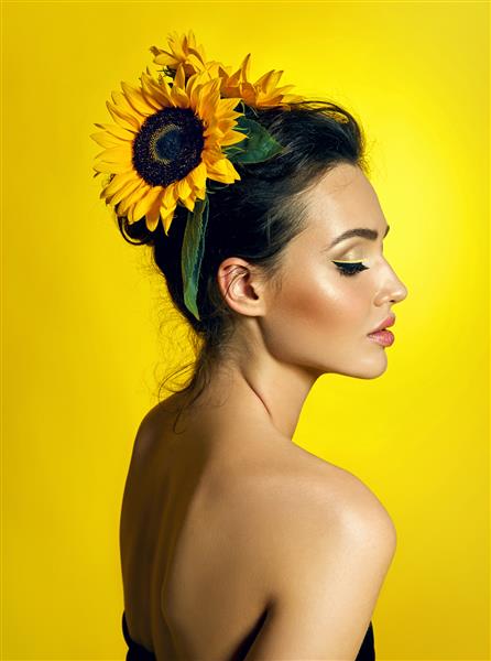 یک دختر جوان با موهای تیره زیبا با پوستی کاملاً صاف و نیم چرخش روی پس زمینه زرد هزینه دارد گل آفتابگردان به شکل تزئینات در مو