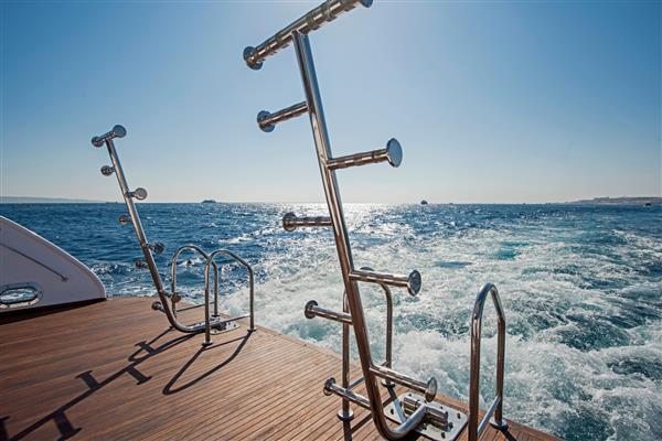 نردبان های فلزی روی عرشه چوب ساج پشتی یک قایق بادبانی موتوری لوکس که در اقیانوس گرمسیری با پس زمینه آسمان ابری قایقرانی می کند