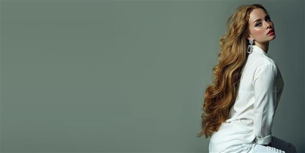 دختر جوان و زیبای چشم آبی با موهای بلند مجلل قرمز موج دار که در استودیو با لباس های سفید ژست گرفته است