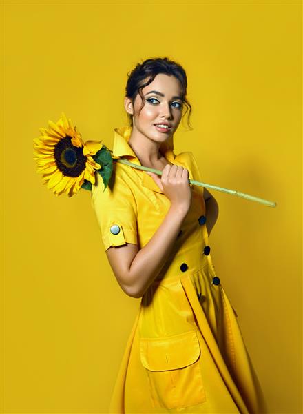 دختری با لباس زرد یک گل آفتابگردان بزرگ در دست دارد و با بازیگوشی لبخند می زند