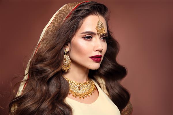 زن زیبا با مدل موهای موج دار بلند و آرایش روشن مدل سبزه هندو جوان با ست جواهرات طلایی کوندان جدا شده روی پس‌زمینه قهوه‌ای استودیو