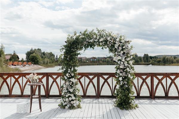 طاق عروسی تزئین شده با گل های سفید در پس زمینه رودخانه بیرون محل برگزاری مراسم عروسی