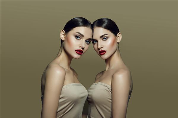 دو زن جوان زیبا با موهای تیره جمع‌شده با پوستی عالی و لب‌های قرمز روشن در تاپ‌های بژ روی پس‌زمینه بژ در استودیو