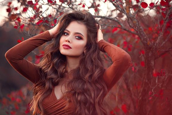 پرتره پاییزی در فضای باز از زن جوان زیبا با آرایش و موهای موج دار بلند که روی پارک پاییزی قرمز ژست گرفته است