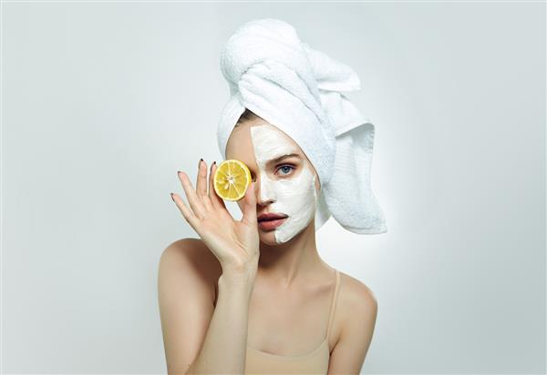 زن جوانی زیبا با حوله ای سفید روی سر و ماسک کرم آرایشی روی صورتش تکه ای لیمو در دست دارد زیبایی شناسی