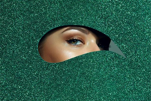 دختری با چشمان سبز زیبا با سایه های روشن روی پلک هایش از سوراخ قطره اشکی که از کاغذ براق سبز ساخته شده است نگاه می کند