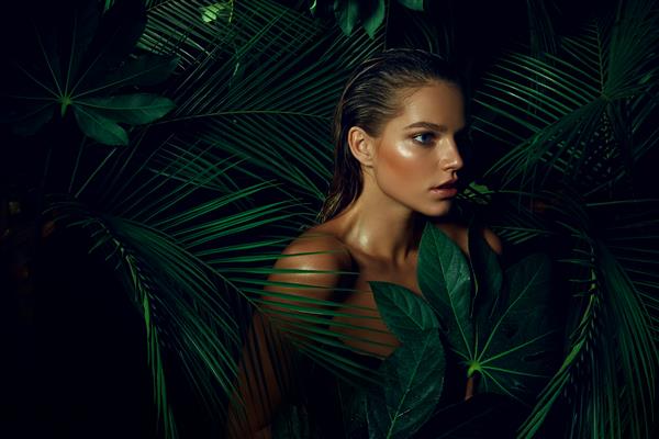 یک زن جوان برنزه زیبا با آرایش طبیعی و موهای خیس در جنگل در میان گیاهان عجیب و غریب ایستاده است گیاهان گرمسیری طبیعت