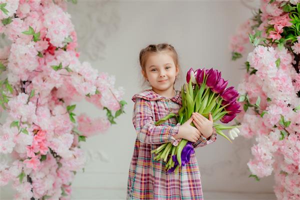دختر کوچولوی دوست داشتنی با لباسی که دسته گل لاله در دست دارد در یک استودیوی عکاسی روشن