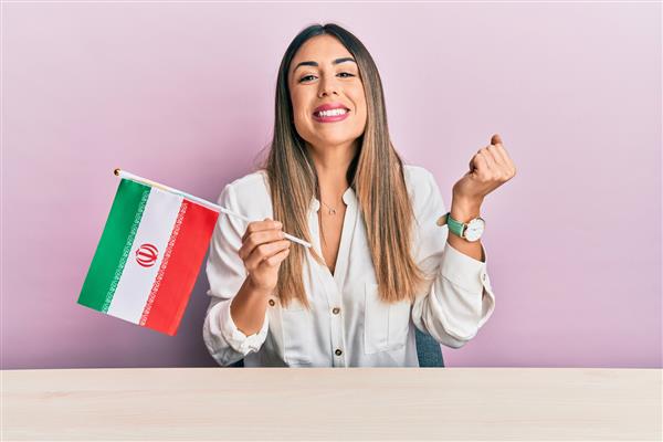زن جوان اسپانیایی تبار پرچم ایران را در دست دارد و روی میز نشسته و با غرور فریاد می زند پیروزی و موفقیت را جشن می گیرد و با بازوی بلند شده بسیار هیجان زده است