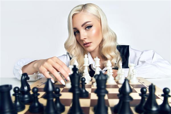 زن بلوند جوان زیبا با آرایش مد قدیمی شطرنج روی پس زمینه سفید بازی می کند مفهوم تبلیغات استراتژی و برنامه ریزی کسب و کار تبلیغات لباس و لوازم آرایش