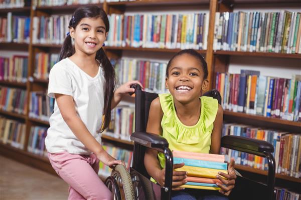 پرتره دانش آموز خندان روی صندلی چرخدار که کتاب در کتابخانه مدرسه در دست دارد