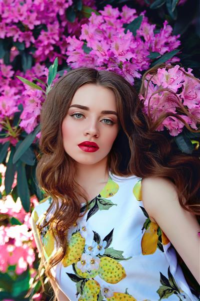 دختری زیبا در لباسی روشن با موهای روان در پس‌زمینه درختچه‌هایی با گل‌های صورتی بزرگ و برگ‌های بزرگ