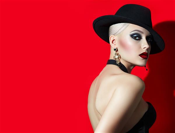دختر شیک پوش زیبا با سنگ موهای بلوند در کلاه سیاه با لب های قرمز چشم های دودی دختر در استودیو در پس زمینه قرمز عکاسی تبلیغات لوازم آرایشی و بهداشتی Photostudio جواهرات جواهر