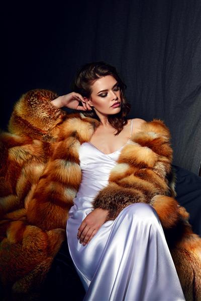 دختری زیبا با موهای قهوه ای بلند و ترکیبی از لباس ساتن سفید در کت خز نشسته است ملکه برفی