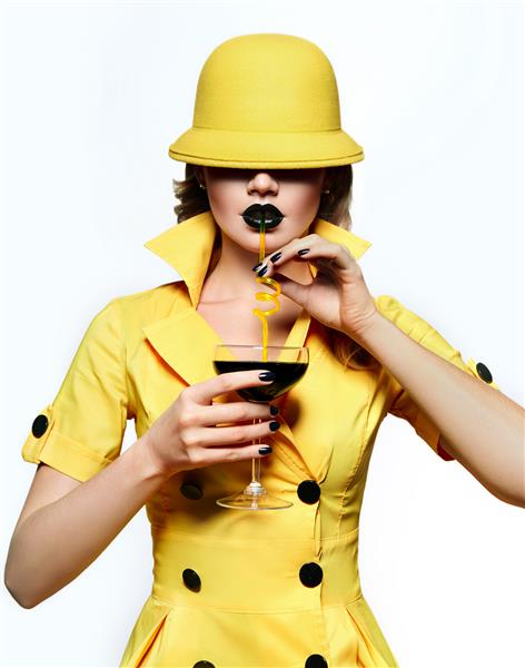 یک دختر جوان زیبا با کلاه زرد و لباس زرد لیوانی با یک کوکتل سیاه و نوشیدنی از یک نی در دست گرفته است