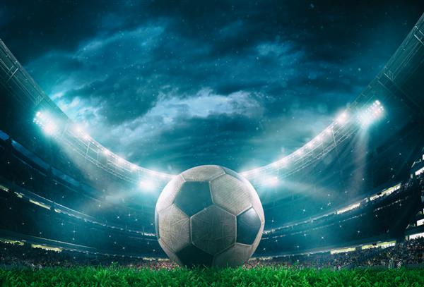 نمای نزدیک از یک توپ فوتبال در مرکز استادیوم که توسط چراغ های جلو روشن شده است