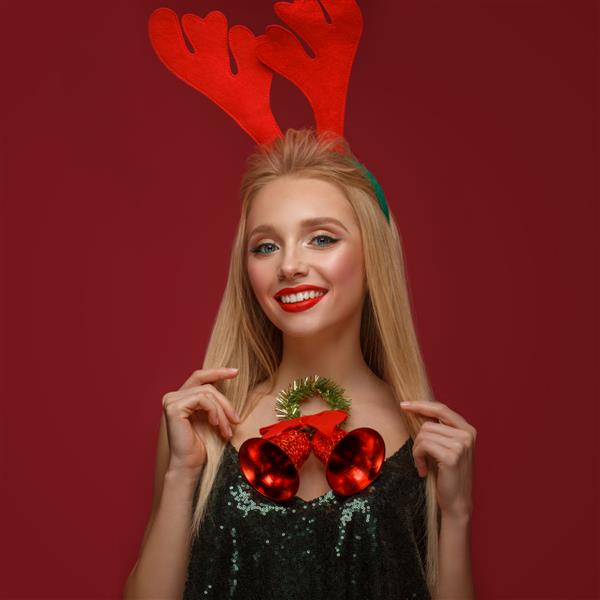 دختر بلوند زیبا در تصویر سال جدید با زنگ های کریسمس به دور گردن و شاخ گوزن روی سرش صورت زیبایی با آرایش جشن عکس گرفته شده در استودیو