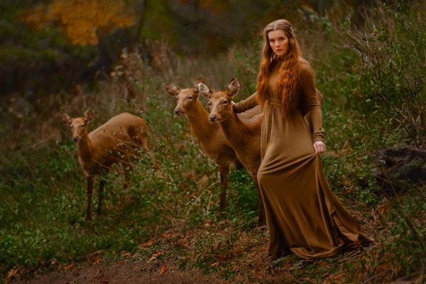 دختر مو قرمز با لباس بلند در جنگل با آهو