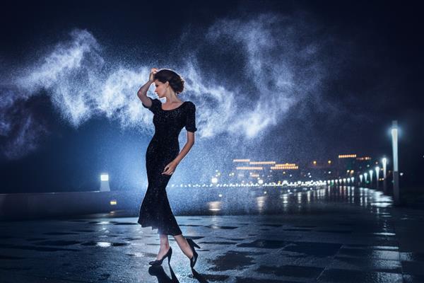 زن زیبا با لباس مشکی که روی پس‌زمینه چراغ‌های شهر ژست گرفته است