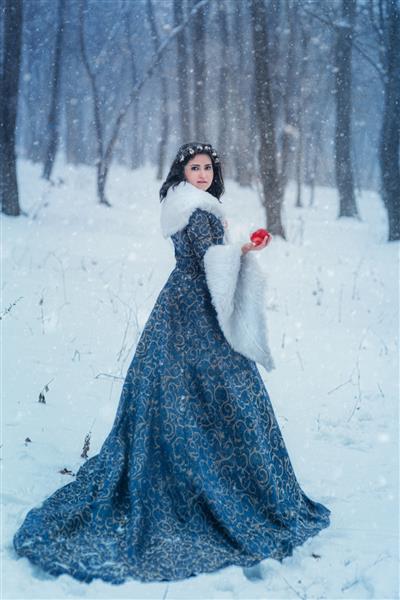 پرتره زن جوانی که کت آبی پوشیده است او در حال قدم زدن در جنگل زمستانی است و سبدی با سیب در دست دارد