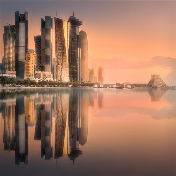 خط افق خلیج غربی و مرکز شهر دوحه در هنگام غروب خورشید با انعکاس ساختمان‌ها روی آب قطر